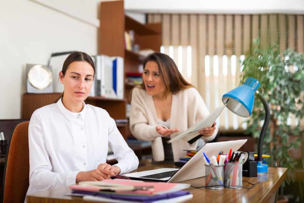 Angry business woman criticizing sad frustrated female employee sitting at office desk at work