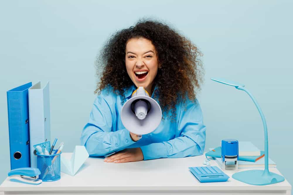 Young excited employee business woman wears casual shirt sit work at white office desk scream in megaphone announces discounts sale Hurry up isolated on plain pastel blue background studio portrait