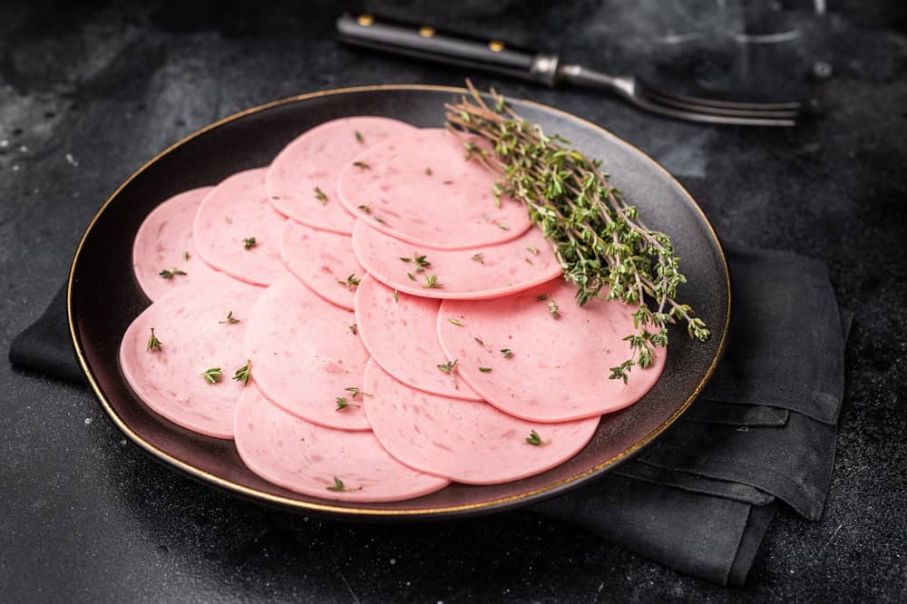 Sliced pink lunch meat (bologna mortadella) with fresh thyme on a black plate. Delicious cold cut and charcuterie concept on a dark background