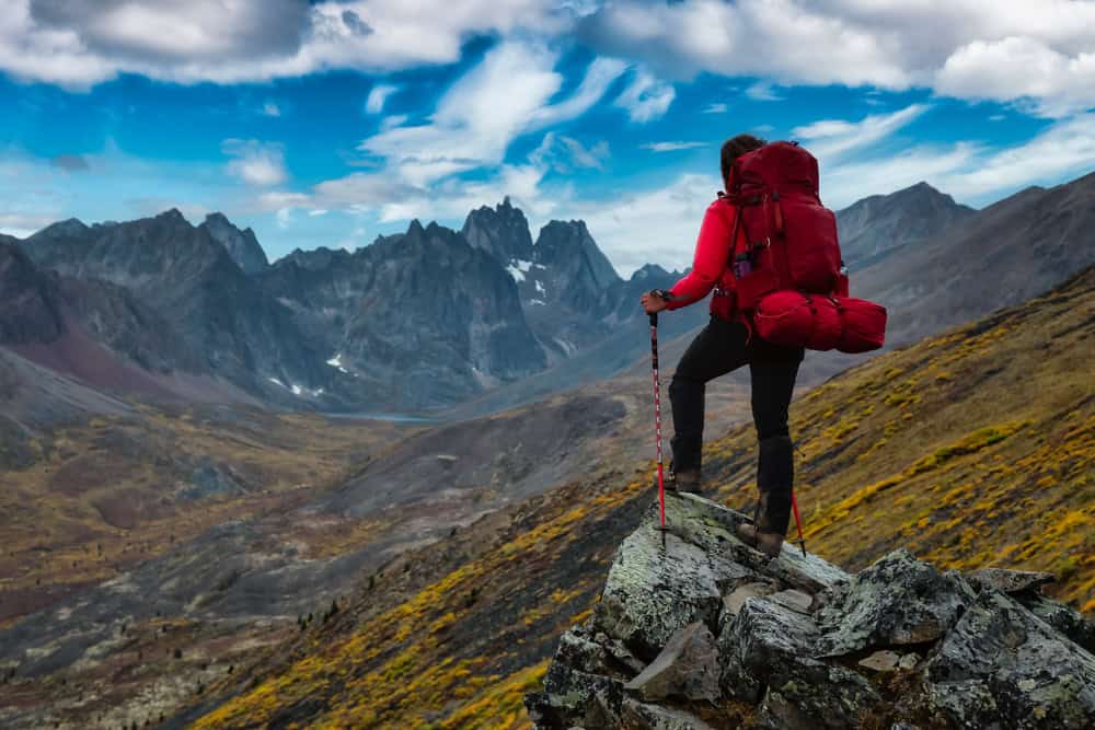 Woman Standing on Rocks looking at Scenic Mountain Peaks and Valley during Fall in Canadian Nature. Colorful Cloudy Sky Artistic Render. Landscape in Tombstone Territorial Park, Yukon, Canada.